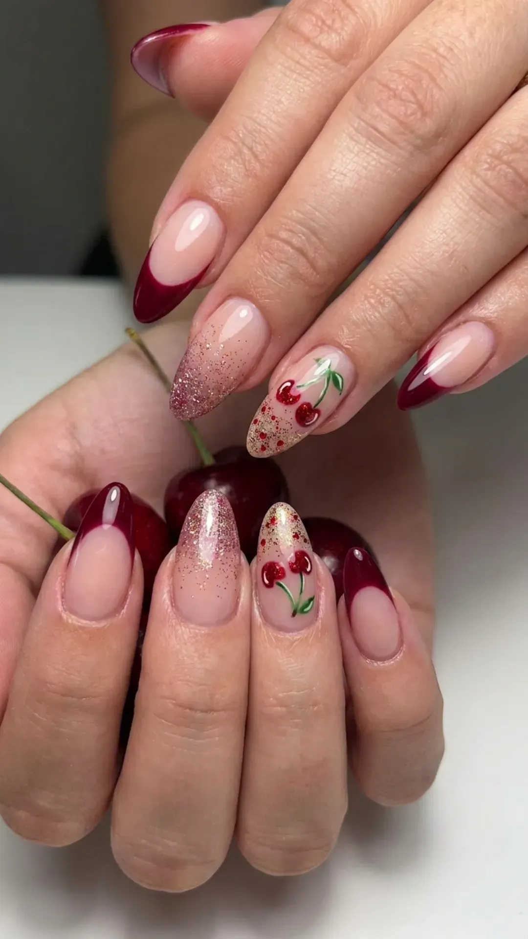 Deep red French tip nails accented with cherry designs and glitter for a flirty and fun manicure.