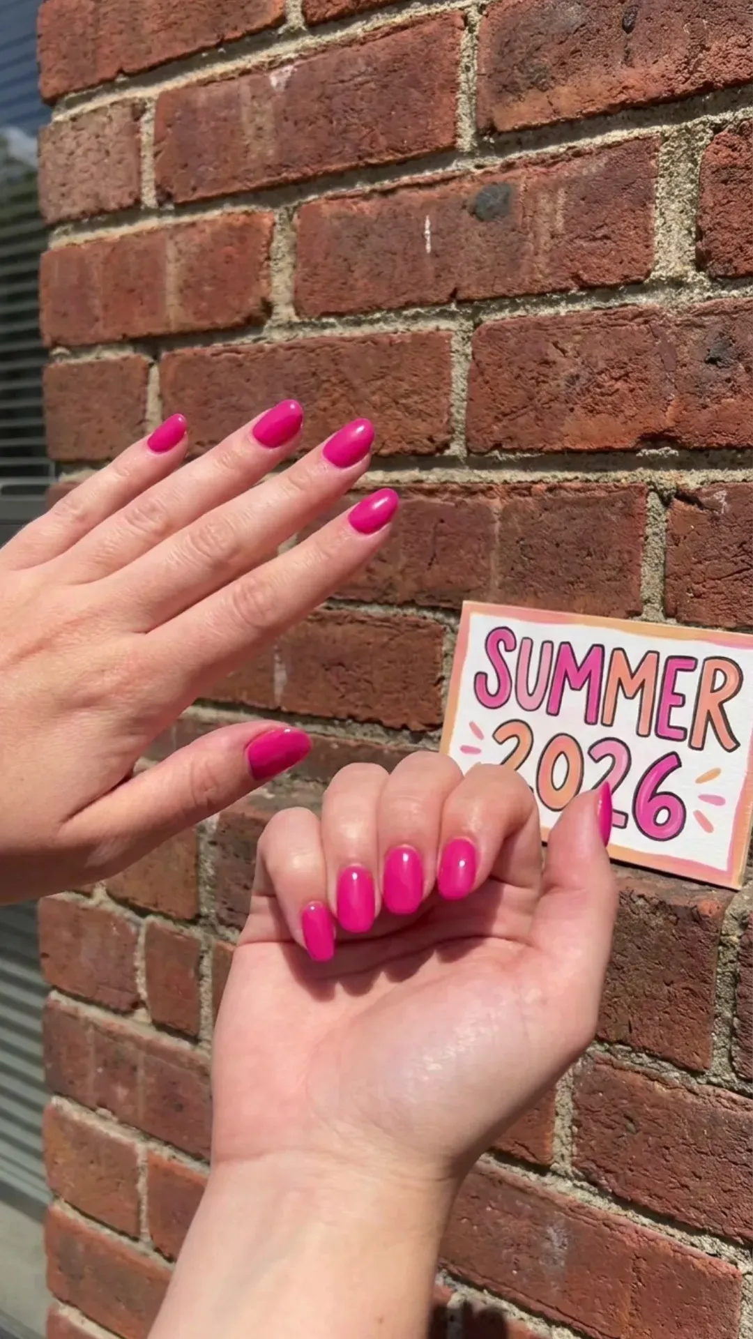 Two hands with hot pink nails against a brick wall, emphasizing a vibrant and lively color perfect for summer 2026.
