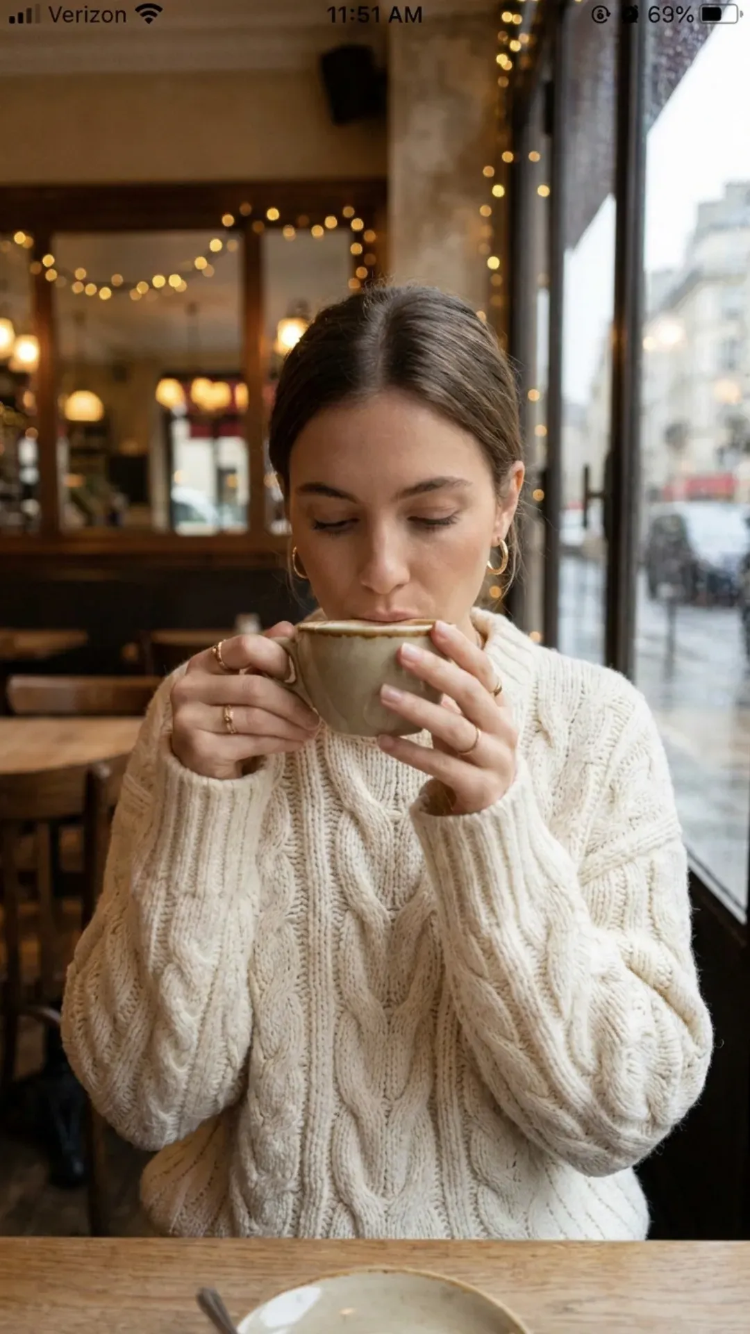 A woman in a cozy cafe wearing an oversized cream cable knit sweater and gold hoop earrings while holding a coffee cup.