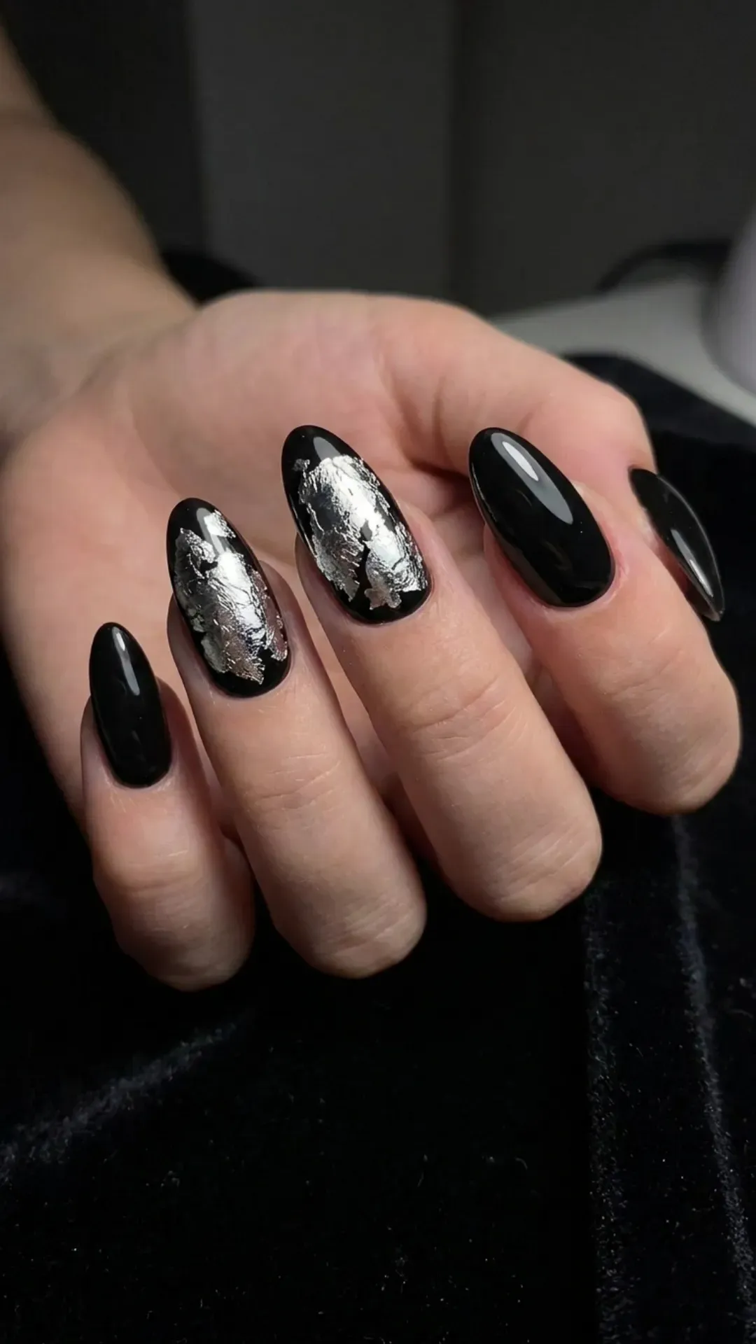 Black almond nails accented with bold silver foil pieces on selective fingers, creating a dramatic and luxurious contrast that's eye-catching.