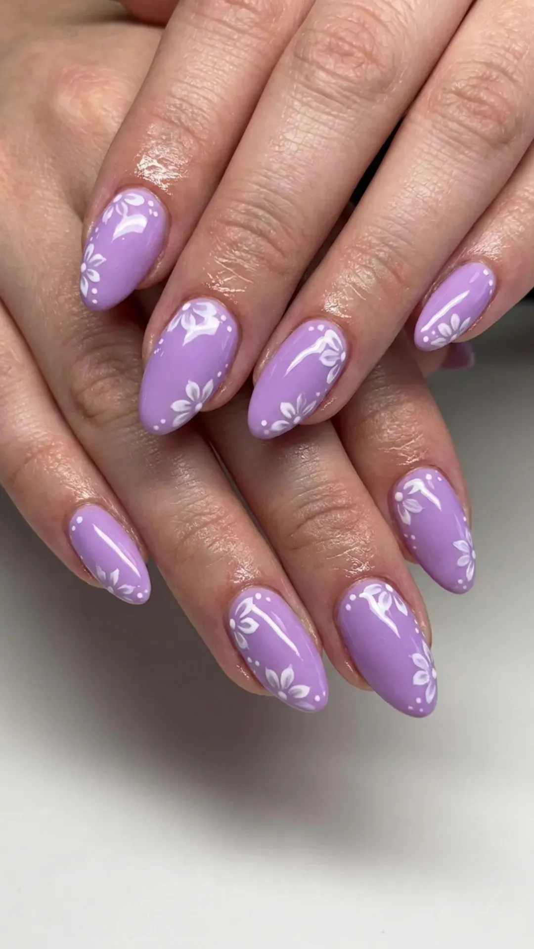 Lavender nails with a glossy finish and white flower designs, accented with small white dots near the cuticles