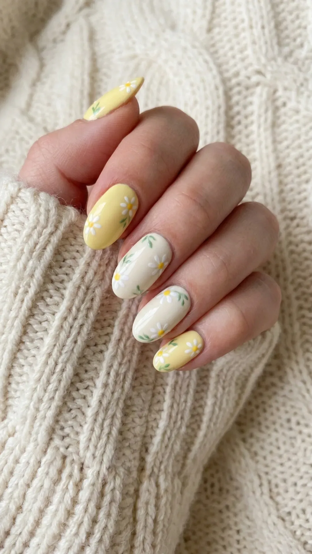 Oval nails painted in alternating shades of pastel yellow and cream, decorated with small daisy patterns and green leaves