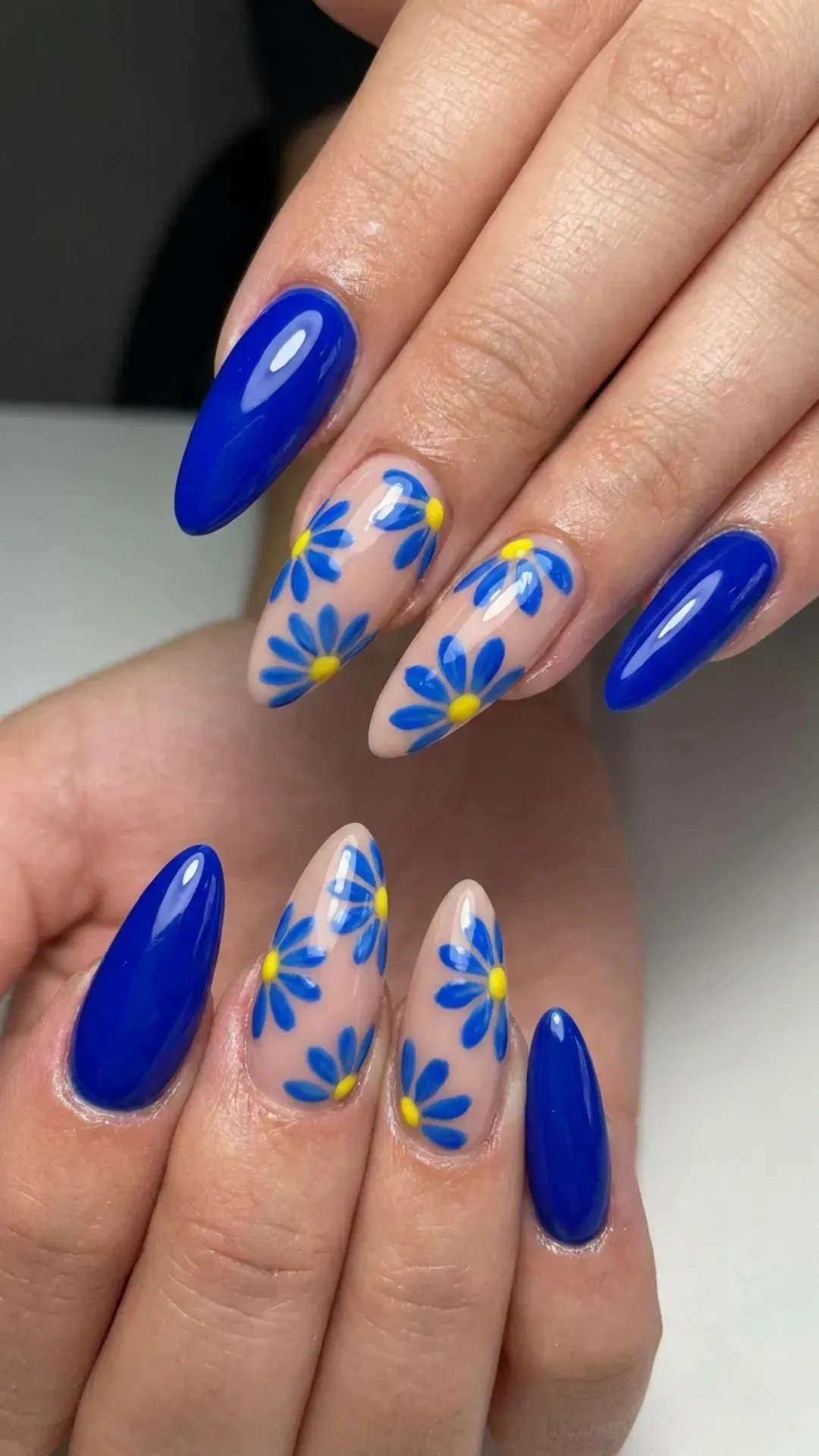Long almond nails with a mix of blue daisy designs and solid blue color on a nude base, giving a bold statement look