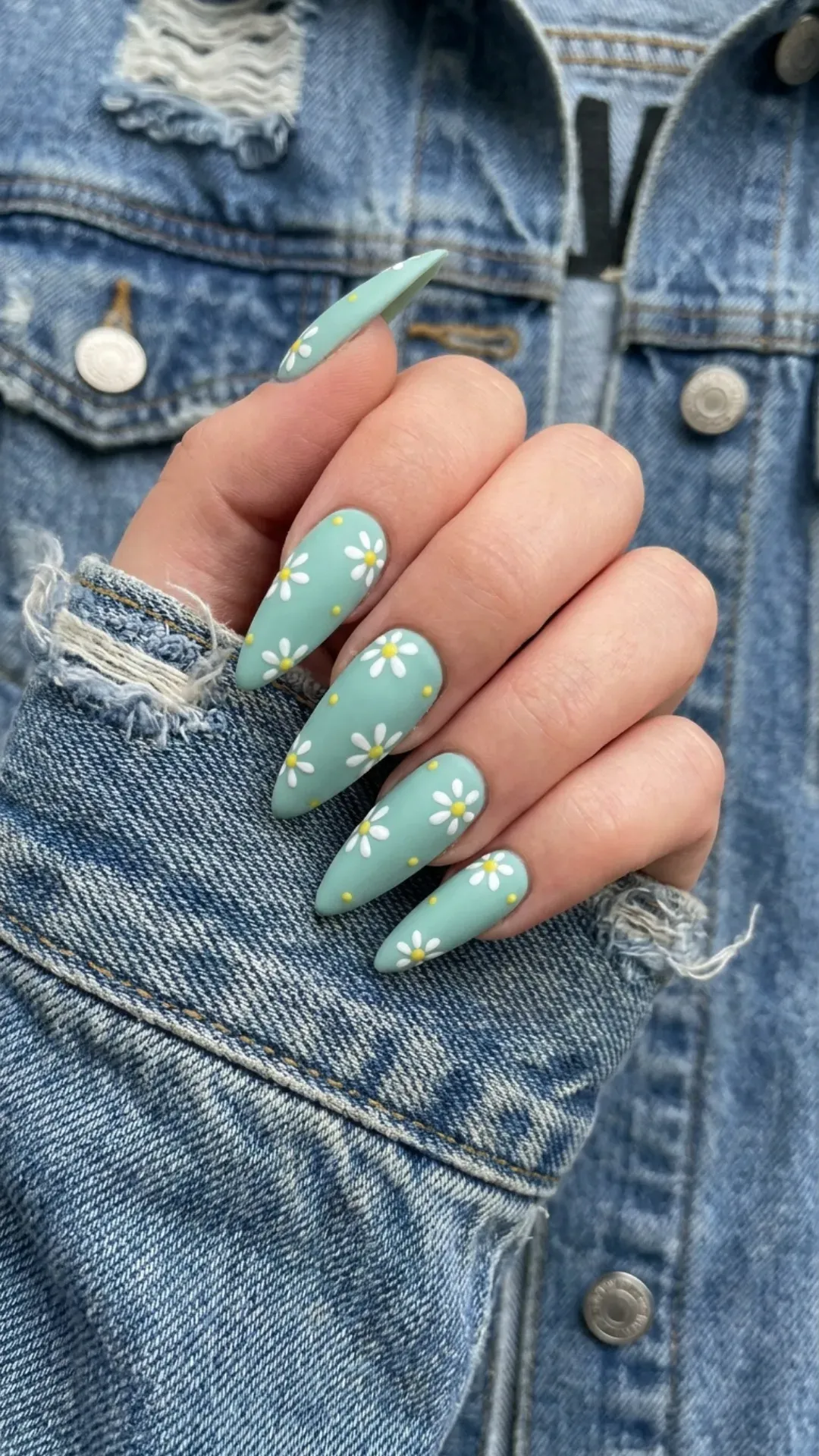 Pointed long nails painted in a soft seafoam green, decorated with white daisies and tiny yellow centers, contrasting with denim