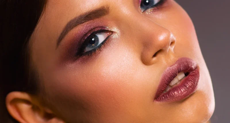 Close-up of a woman's face showcasing shimmering eyeshadow, full lashes, and glossy mauve lipstick, highlighting a glamorous makeup look