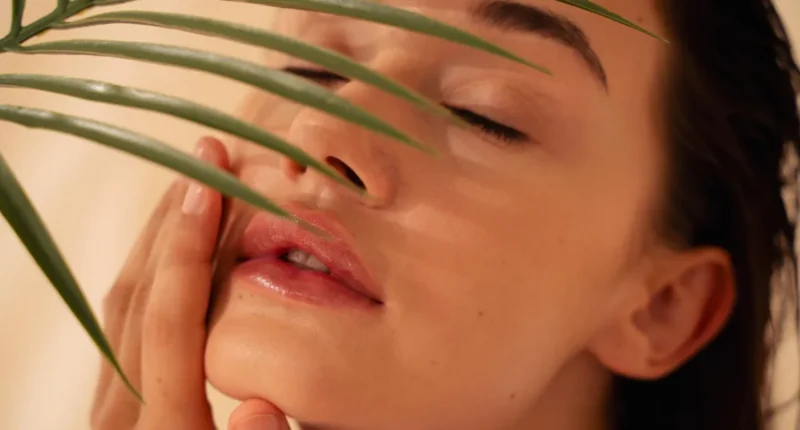 A close-up of a woman with a serene expression, having a green leafy plant brushed over her face, depicting a botanical skincare treatment.