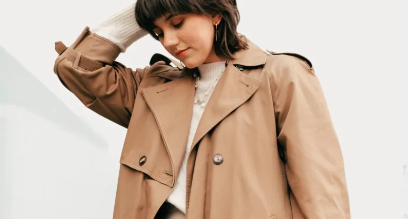 Woman in a chic beige trench coat touching her hair, epitomizing classic winter fashion trends for 2024 with a street-style vibe.