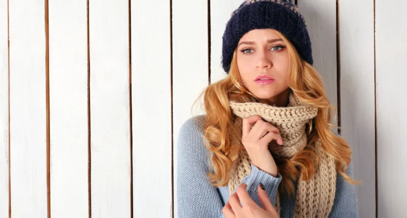 A thoughtful woman in cozy winter attire, featuring a chunky knit scarf, a woolen beanie, and a blue sweater, set against a crisp white wooden backdrop, embodying the warmth and style of the season