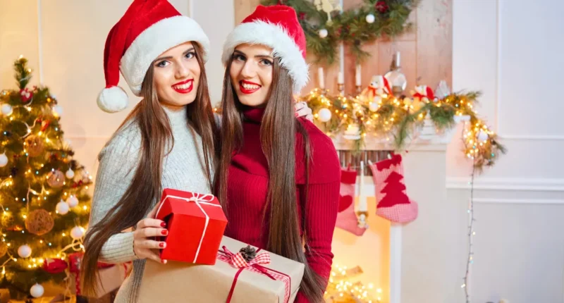 Two joyful women in festive Christmas attire holding presents, wearing Santa hats, with a beautifully decorated Christmas tree in the background.