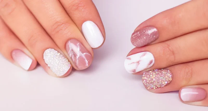 Assorted festive nail designs featuring a white base with varied accents: one with white pearl beads, another with marble effect, one sparkling with pink glitter, and a couple with a soft pink ombre, perfect for holiday celebrations or a chic winter look