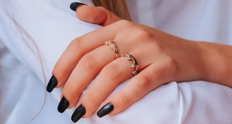 A woman's hand with elegant gold rings, featuring long, almond-shaped nails painted in a glossy black polish.