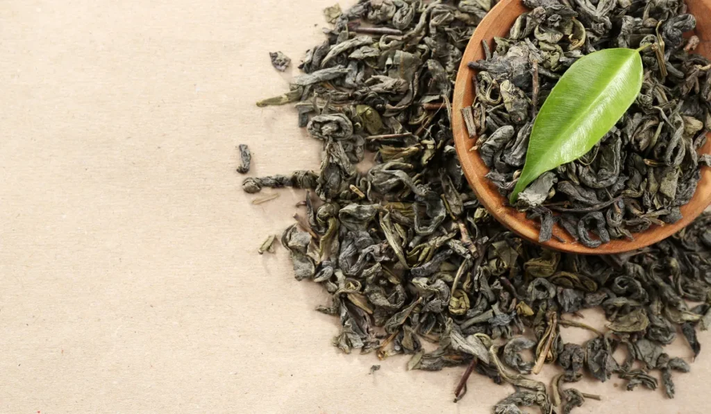 Close-up of a green tea leaf on top of dried tea leaves in a wooden bowl, highlighting natural antioxidants beneficial for health and skincare.
