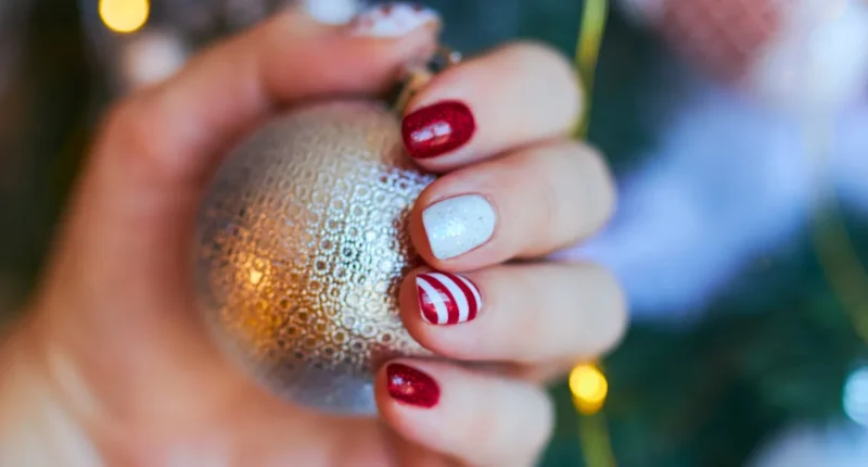 Hand holding a festive bauble showcases a Christmas gel nail manicure with sparkling red and silver glitter nails and a feature nail with candy cane stripes, embodying the cheerful and cozy holiday spirit of Christmas 2023.