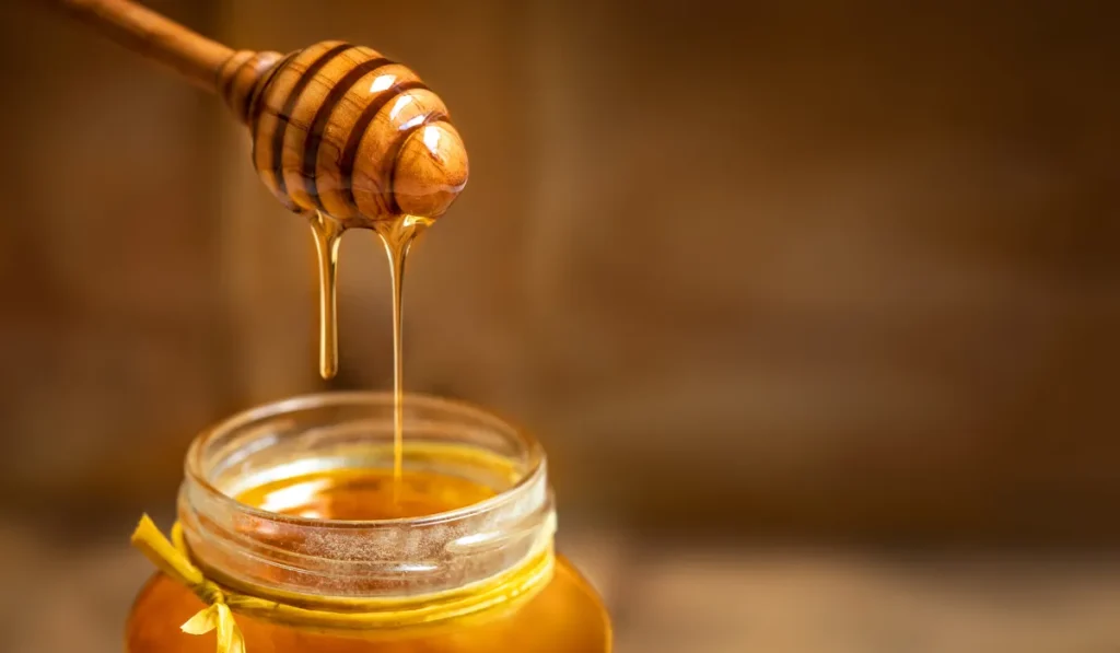 A close-up of a wooden honey dipper dripping golden honey into a jar, representing natural ingredients for skincare or healthy eating.
