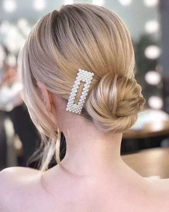 A sleek and elegant blonde low bun hairstyle, adorned with a large pearl barrette, perfect for bridal or formal events.
