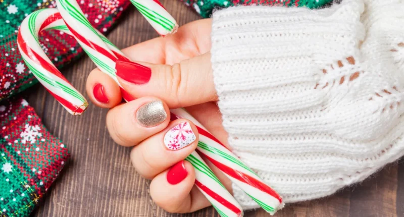 A hand displaying festive Christmas nail art, with glossy red polish on most nails, one accent nail in glittering gold, and another featuring a delicate snowflake design on a white base. The nails complement the seasonal theme, complete with traditional candy canes held in the hand, and are set against a backdrop of cozy winter knitwear and festive decorations.