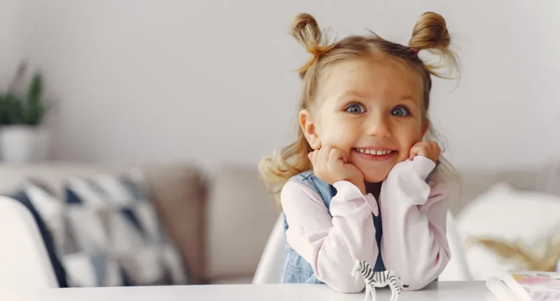 A cheerful young girl with blonde hair styled into playful high buns, blue eyes, and a wide smile, sitting at a white table in a bright, cozy room.