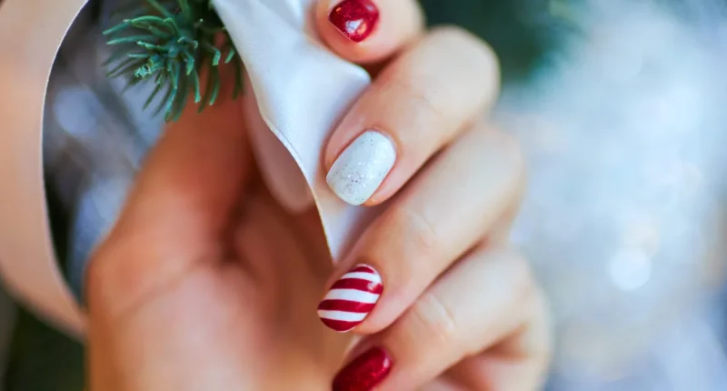 A close-up of a hand showcasing simple Christmas-themed nail art, featuring alternating candy cane stripes and delicate snowflakes on a white polish base