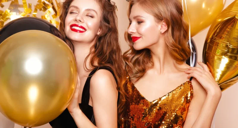 Two smiling women in glamorous party outfits with gold balloons, one wearing a sequin dress and the other in a black top with a red lipstick.