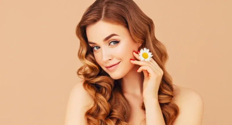 A woman with flowing chestnut hair and a subtle smile, holding a white daisy flower to her cheek, exemplifying natural beauty against a soft beige background."