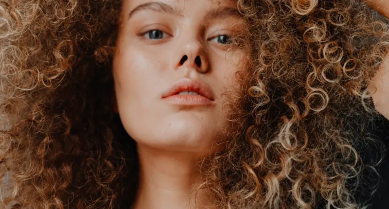 Close-up of a woman with voluminous naturally curly hair, showcasing a variety of tight curls and a rich, sun-kissed ombre color, exuding confidence and a carefree spirit.