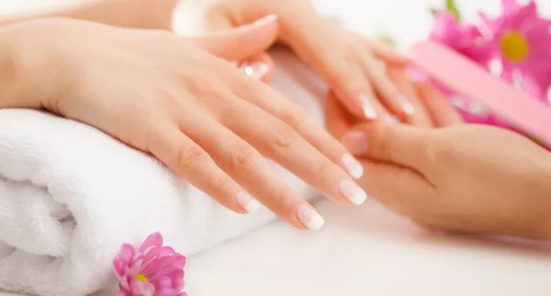 A person's hands gently resting on a soft white towel with manicured nails featuring a classic French tip design, accompanied by pink blossoms emphasizing a pampered and delicate spa atmosphere.