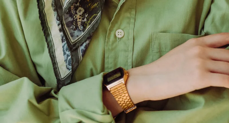 Close-up of a stylish sage green shirt with intricate scarf detail at the neckline, complemented by a vintage gold digital watch on a slender wrist.