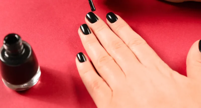 Close-up image capturing the precise application of glossy black nail polish on short, neatly manicured nails over a vibrant red background, highlighting the beauty of simple yet bold nail color choices.