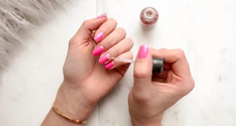 applying-sparkle-top-coat-pink-nails Alt Text: A close-up image capturing the moment a clear sparkle top coat is being applied to vibrant pink nails, the brush leaving a trail of shimmering glitter, perfect for adding a festive touch to a bold manicure. The process is set against a cozy, white furry background, emphasizing the warmth and comfort of the winter season.