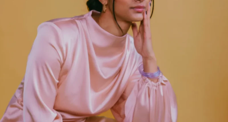 A close-up of a person against a yellow background, wearing a blush pink satin top with gathered sleeves, adorned with subtle gold earrings and a set of thin purple bangle bracelets