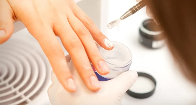 A person carefully applies a clear liquid to the nails over a white dip powder container, preparing for the dip powder nail process, with a blurred fan in the background indicating a dust-free environment