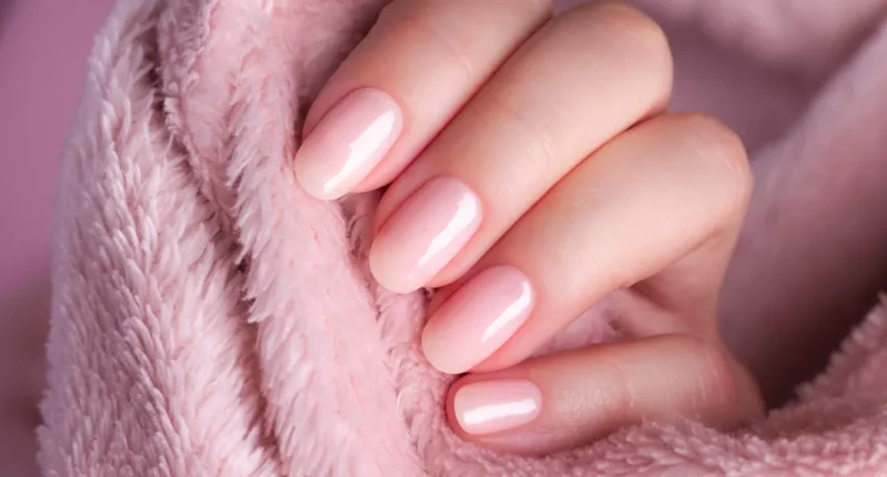 Close-up of a hand wrapped in a plush pink fabric, showcasing a set of impeccably manicured nails painted in a soft nude pink shade that exudes warmth and comfort, perfect for a cozy day in.