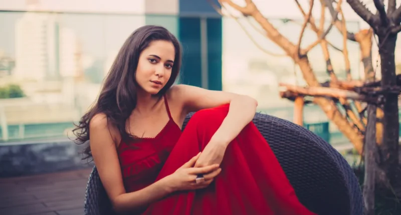 Elegant woman sitting in a wicker chair outdoors, wearing a vibrant red Valentine's Day dress with delicate lace detailing and a flowing skirt, perfect for a romantic evening