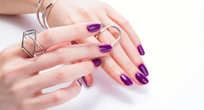 A hand with oval-shaped nails lacquered in a vibrant glossy purple shade, accessorized with modern geometric rings, one shaped as an abstract polygon and the other as a simple open band, set against a bright white background.