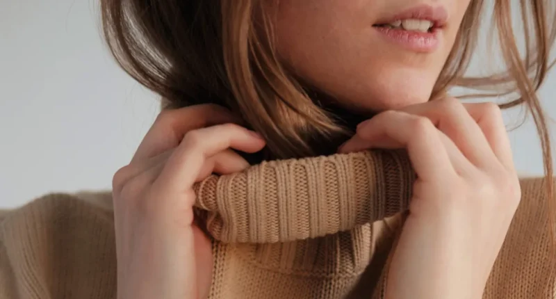 A close-up of a woman adjusting a high-neck, tan knit sweater, focusing on the soft texture and simple elegance of the garment, perfect for a casual yet sophisticated look