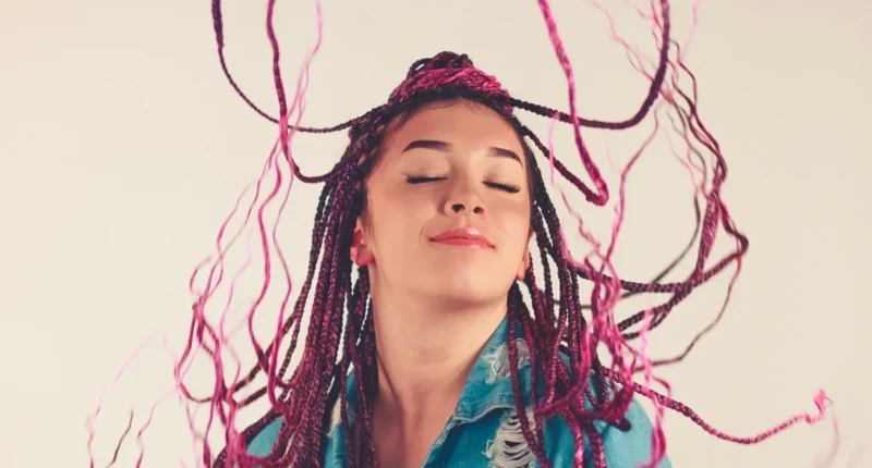 A dynamic image capturing a woman with a joyful expression, her hair in black and pink braids flying around as she likely moves in a dance, against a soft background.