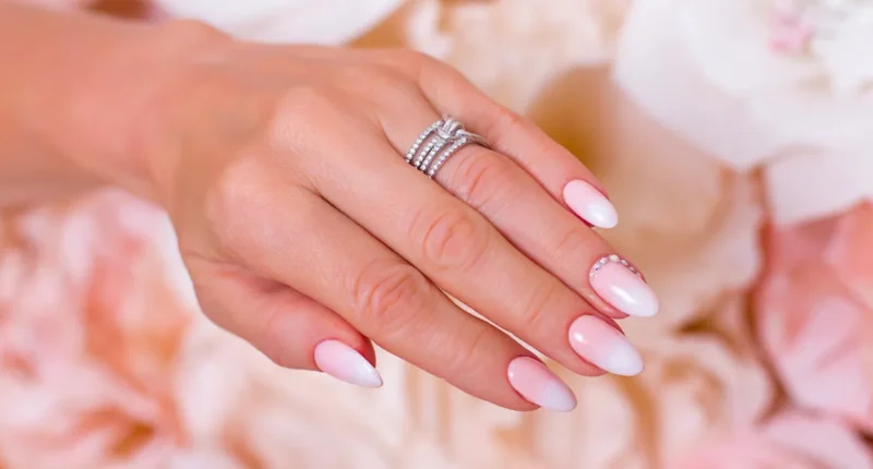 Elegant soft blush ombre nails with a delicate white gradient, adorned with a subtle rhinestone accent on the ring finger, set against a romantic floral background, perfect for a bridal look or a sophisticated spring/summer occasion.