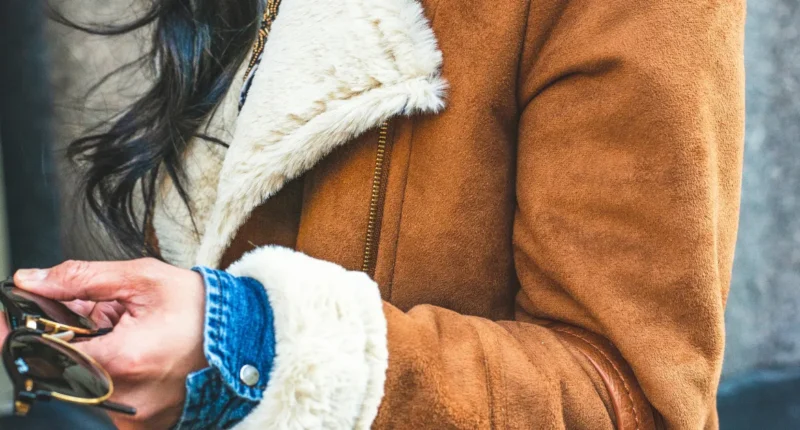 Close-up of a woman holding sunglasses, wearing a trendy brown suede jacket with a cozy white faux fur collar and a blue denim shirt underneath, a stylish detail for a winter fashion ensemble.