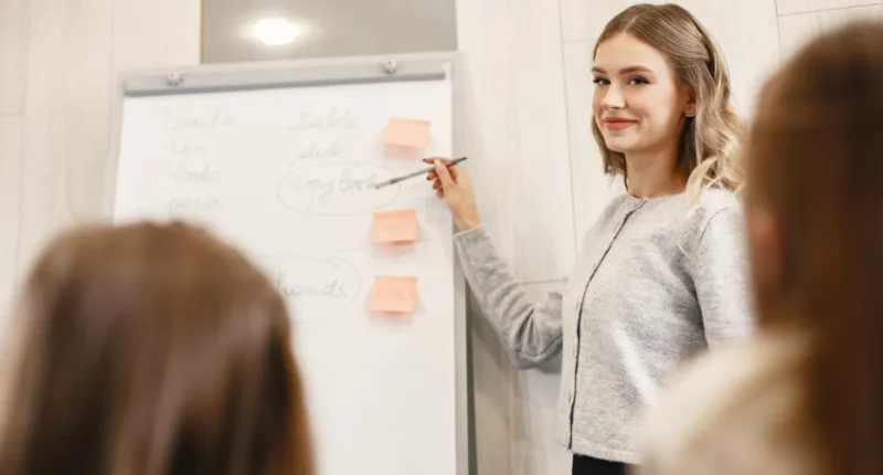 A modern teacher in a smart grey sweater and subtle makeup engaging with students during a classroom presentation, standing beside a whiteboard adorned with sticky notes, exuding a professional and approachable demeanor suitable for a winter school day.