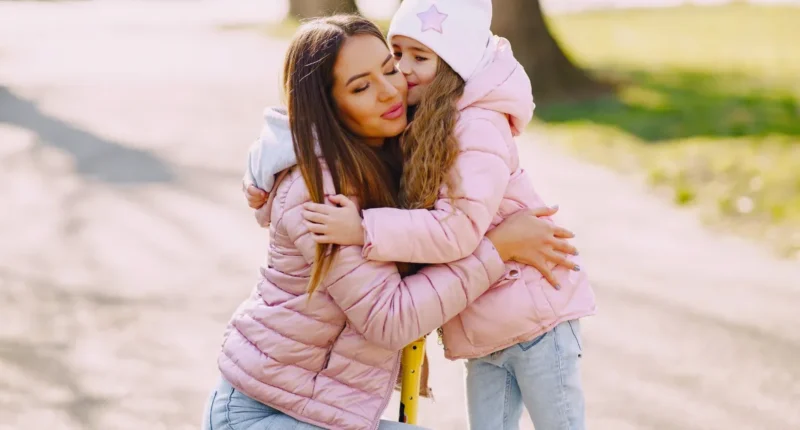 A tender moment captured as a mother and daughter, both wearing matching pastel pink puffer jackets and denim jeans, embrace warmly on a sunny winter day, with expressions of love and affection