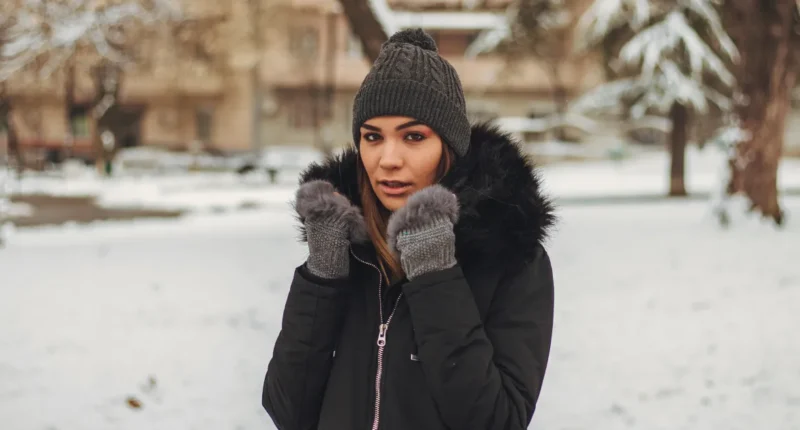 A stylish woman outdoors in winter wearing a black insulated jacket with a fur-lined hood, knit beanie, and grey gloves, ready for a cold urban adventure with a backdrop of snow-covered trees.