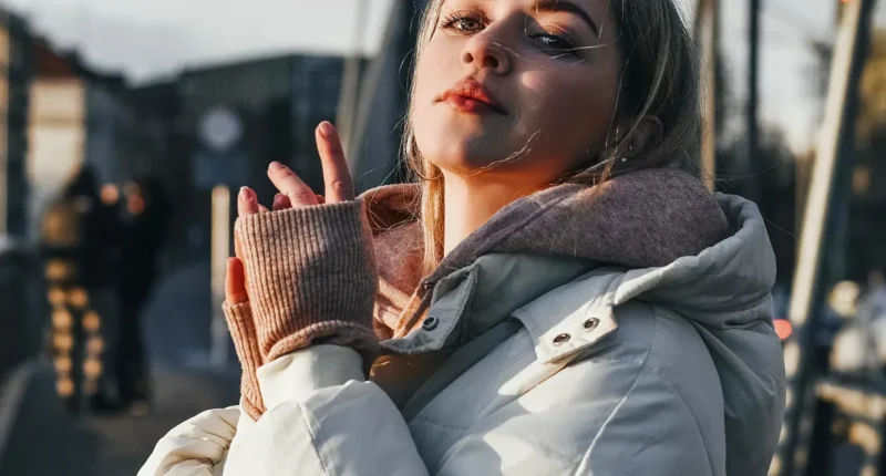 A woman basking in the winter sunlight on a bustling city street, showcasing a modern winter fashion look with a cozy beige turtleneck, warm fingerless gloves, and a chic ivory puffer jacket, perfectly capturing the essence of stylish urban life in the colder months.