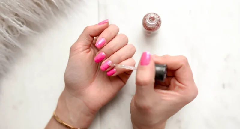 Close-up of hands applying a shimmering sparkle over vibrant pink polish, capturing the process of creating a dazzling Valentine's Day nail design.