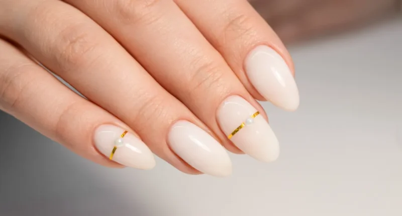 Sophisticated white almond nails with a minimalist design, featuring a single pearl and gold stripe accents, suitable for elegant bridal looks or formal events.