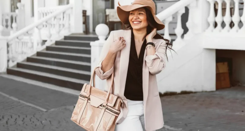 Joyful woman in spring attire wearing a wide-brimmed hat, nude blazer, and carrying a large tote bag, laughing on the steps of a white building, embodying the light-hearted spirit of spring