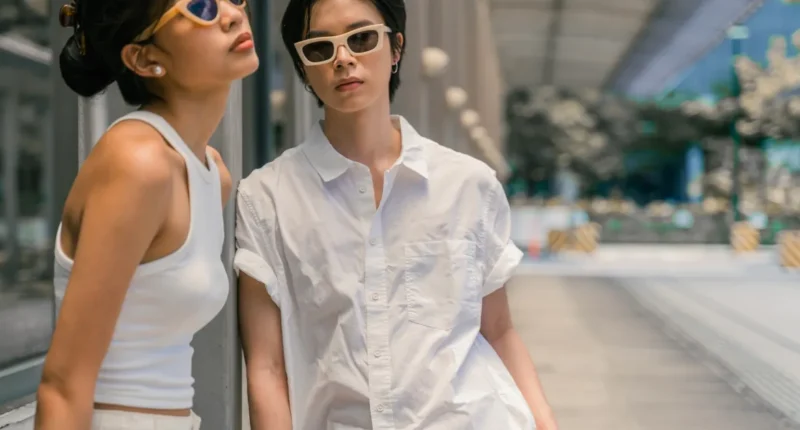 Two fashion-savvy individuals in urban streetwear, with one donning a white sleeveless top and the other in an oversized white button-down, both with trendy sunglasses, embodying effortless city style