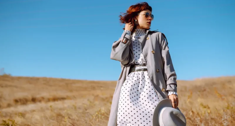 A stylish woman outdoors wearing a white and black polka dot dress beneath a classic grey trench coat, complete with a silver belt and matching accessories for a chic springtime outfit.