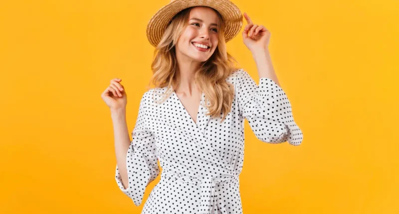 A smiling woman in a classic white and black polka-dot wrap dress with a v-neckline and tie waist, complemented by a straw hat, encapsulating a timeless summer style for picnics or seaside walks.