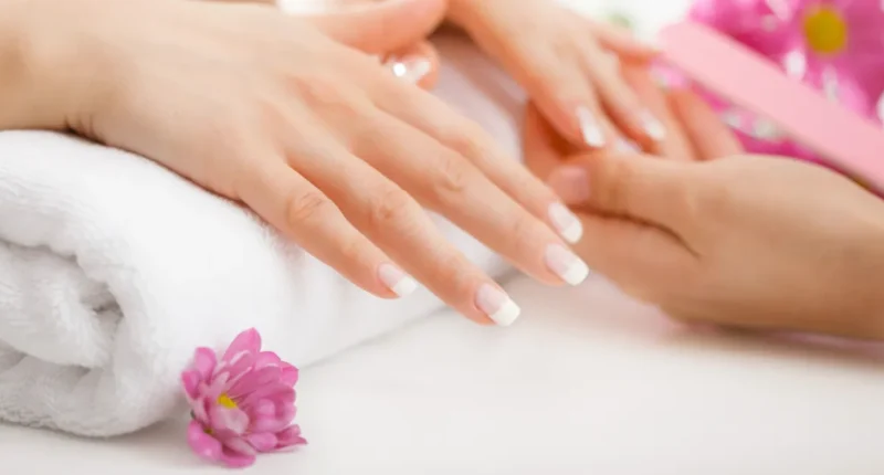 Detailed view of a French manicure being applied to a woman's nails, featuring a hand resting on a towel with a pink flower, showcasing simple at-home summer nail care techniques