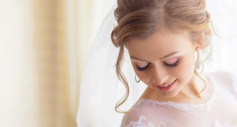 Romantic bridal updo featuring soft curls and a delicate side cascade, complemented by subtle makeup and elegant earrings, perfect for a dreamy and graceful wedding day appearance.